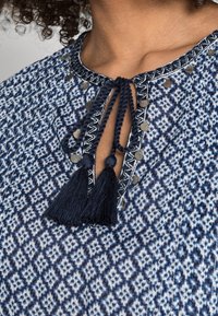 Blusa blu e bianca con un motivo a fantasia, chiusura con nastro a frange, dettagli metallici e tessuto testurizzato con un design geometrico.