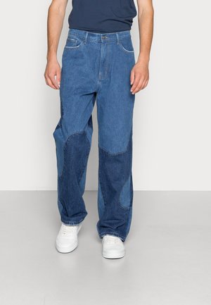 Homme portant un jean en denim bleu patchwork ample et des baskets blanches, debout devant un fond uni.