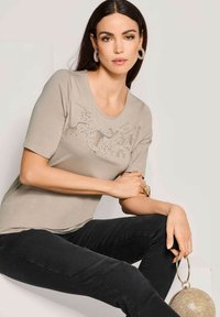 Beige T-Shirt mit kurzen Ärmeln, ausgestattet mit einem strukturierten Design und Verzierungen auf der Vorderseite, kombiniert mit schwarzen Hosen und einer metallischen runden Handtasche.