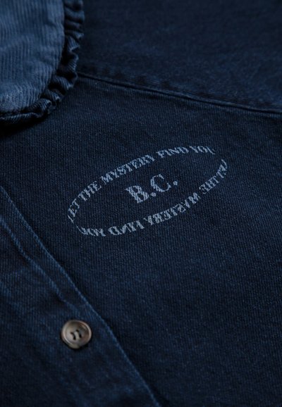 Tissu denim bleu foncé avec un logo ovale imprimé portant la mention "B.C." et la phrase "LET THE MYSTERY FIND YOU" autour, près d’un bouton.