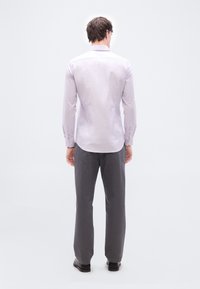 Άνδρας που στέκεται με την πλάτη, φορώντας ανοιχτό μωβ πουκάμισο με μακριά μανίκια, σκούρο γκρι παντελόνι και μαύρα παπούτσια, σε απλό λευκό φόντο.