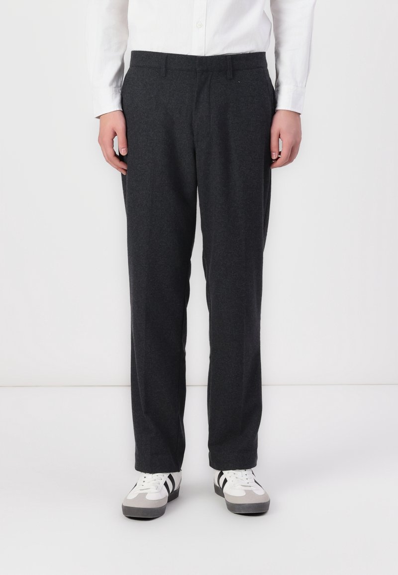 Personne portant un pantalon droit gris foncé, une chemise blanche à manches longues et des baskets blanches avec des détails noirs et gris, debout sur un fond blanc.