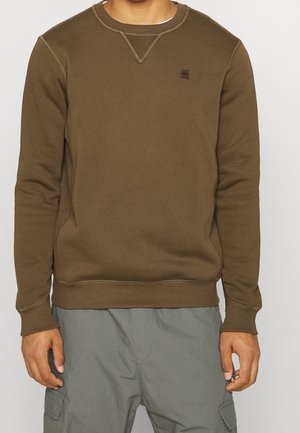 Mand iført en brun crewneck-sweatshirt med kontrastsyning og grå cargobukser, stående mod en ensfarvet baggrund.