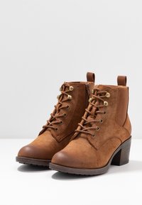 Bottines en daim marron avec un bout arrondi, un laçage à l'avant et une fermeture éclair sur le côté. Présentent des œillets dorés et un talon en bloc en bois.