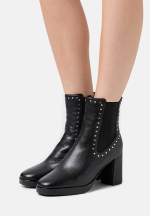 Bottines en cuir noir avec un talon carré haut, des panneaux élastiques sur les côtés et des clous en argent décoratifs le long des bords. Fini texturé.