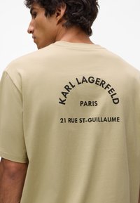 Khaki bavlnené tričko s okrúhlym grafickým potlačou na zadnej strane, ktorá obsahuje tučný čierny text "KARL LAGERFELD," "PARÍŽ," a "21 RUE ST-GUILLAUME."