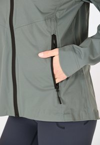 Lichte, groenachtig-grijze ritsjack met een gladde textuur en zwarte accenten, voorzien van twee zijzakken en een getailleerde pasvorm.