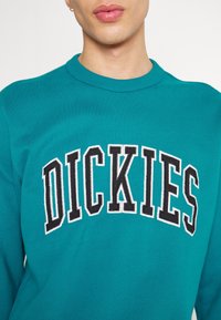 Sweatshirt teal à col rond, arborant le mot "DICKIES" en broderie noire et blanche. Texture douce, coupe décontractée.
