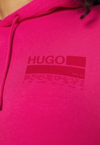 Felpa rosa brillante con un logo "HUGO" di un rosa più scuro e un design pixelato sul petto, dotata di un cordino vicino al collo.