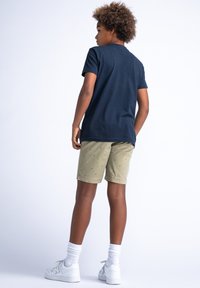 T-shirt di cotone navy, pantaloni corti verde oliva con piccolo motivo a palma, sneaker bianche e calzini bianchi. Vista da dietro, che mostra l'outfit casual.