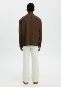Brun oversized skjorta med avslappnad passform, långa ärmar och platt krage, tillsammans med vita vida byxor och bruna skor. Enfärgad bakgrund.
