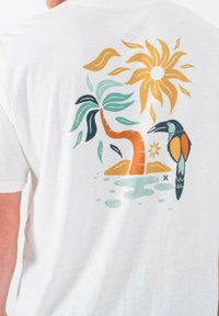 Camiseta blanca con un gráfico de un tucán, una palmera, un sol y elementos acuáticos en colores naranja, verde y azul.