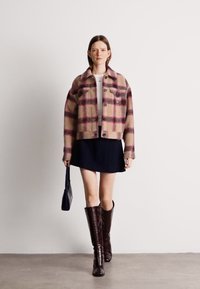 Καφέ και μπορντώ καρό cropped μπουφάν, με μακριά μανίκια, κλείσιμο με κουμπί, συνδυασμένο με μαύρη φούστα και καφέ μπότες μέχρι το γόνατο, κρατώντας μια τσάντα.