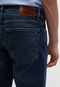 Σκούρο μπλε τζιν με δερμάτινη ετικέτα BOSS στη ζώνη, με κανονικές τσέπες και λογότυπο στην πίσω τσέπη.