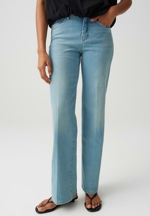 Persona che indossa jeans azzurri a gamba dritta con top nero, una mano in tasca e infradito nere su sfondo semplice.