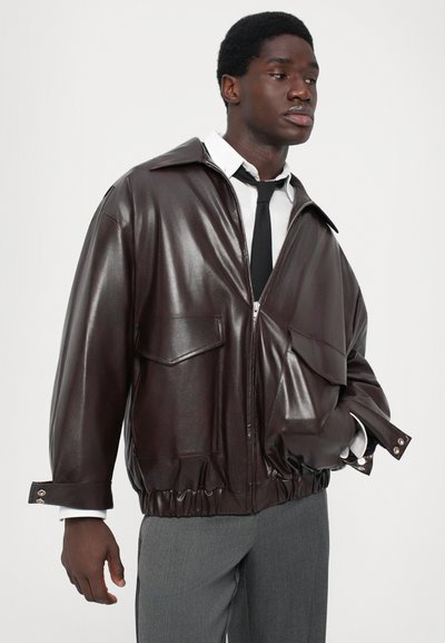 Giacca in pelle marrone con finitura lucida, vestibilità oversize, tasche frontali, orlo elastico e polsini con bottoni a scatto. Indossata con una camicia bianca e una cravatta.