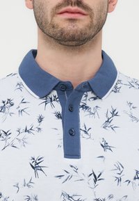 Άνδρας που φοράει πόλο μπλούζα με σχέδιο λουλουδιών σε άσπρο και μπλε, με μονόχρωμο μπλε γιακά και τρία κουμπιά στη λαιμόκοψη, εμφανίζει τον λαιμό και το κάτω μέρος του προσώπου.