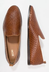 Chaussures slip-on tissées marron en cuir, avec un côté élastique, un bout rond, et une semelle en caoutchouc plate. Nom de la marque "KIOMI" visible à l'intérieur.