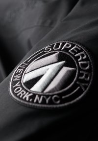Čierna textilná bunda s kruhovým vyšitým logom obsahujúcim nápis "SUPERDRY", geometrický vzor a slová "NEW YORK, NYC."