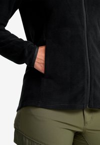 Svart fleecejacka med dragkedja fram, med sidofickor och en mjuk textur. Kombineras med olivgröna byxor som visar del av benet.