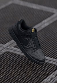 Juodi Nike sportbačiai su tekstūruotu viršumi, lygaus paviršiaus akcentais ir paminkštintu apvadu. Aukso logotipo detalė ant liežuvėlio kontrastuoja su tamsiu dizainu.