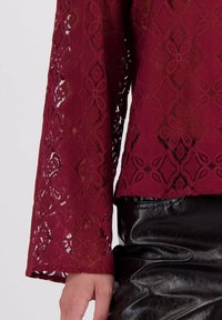 Bordeaux kanten blouse met complexe bloempatronen en lange mouwen, gecombineerd met een glanzende zwarte leren rok die de textuur benadrukt.