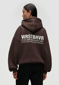 Brown oversized hoodie with a hood, featuring white text on the back: "WRSTBHVR STUDIO (030) BERLIN MON HABIT 0102 CRAFTING AESTHETICS."