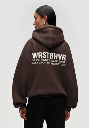 Hoodie surdimensionné marron avec capuche, arborant un texte blanc dans le dos : "WRSTBHVR STUDIO (030) BERLIN MON HABIT 0102 CRAFTING AESTHETICS."