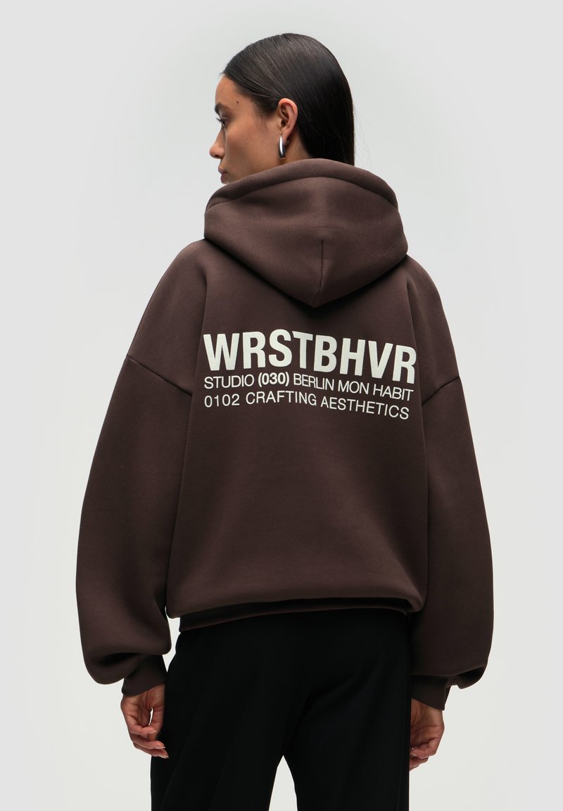 Brown oversized hoodie with a hood, featuring white text on the back: "WRSTBHVR STUDIO (030) BERLIN MON HABIT 0102 CRAFTING AESTHETICS."