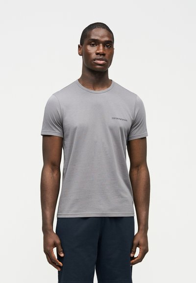 Άνδρας που φοράει εφαρμοστό γκρι ανοιχτό κοντομάνικο μπλουζάκι με μικρό λογότυπο «Emporio Armani», συνδυασμένο με σκουρόχρωμο σορτς, σε μονόχρωμο φόντο.
