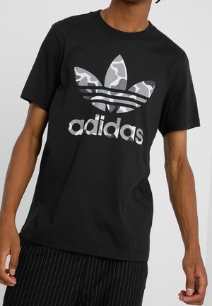 Camiseta de algodón negra con un gran logo de Adidas en tonos de gris, camuflado, y tres franjas horizontales encima del logo.