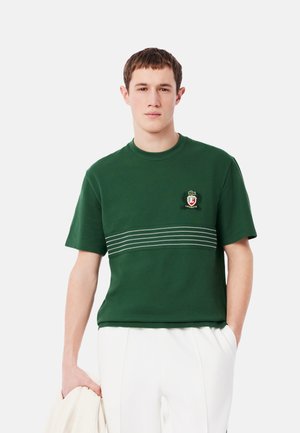 Camiseta de algodón verde con cuello redondo, mangas cortas y un detalle de rayas horizontales con un logo bordado en el pecho.