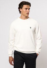 Sweatshirt blanc à manches longues en tissu doux, avec un col rond, des poignets et un ourlet côtelés, et un petit logo noir sur la poitrine gauche.