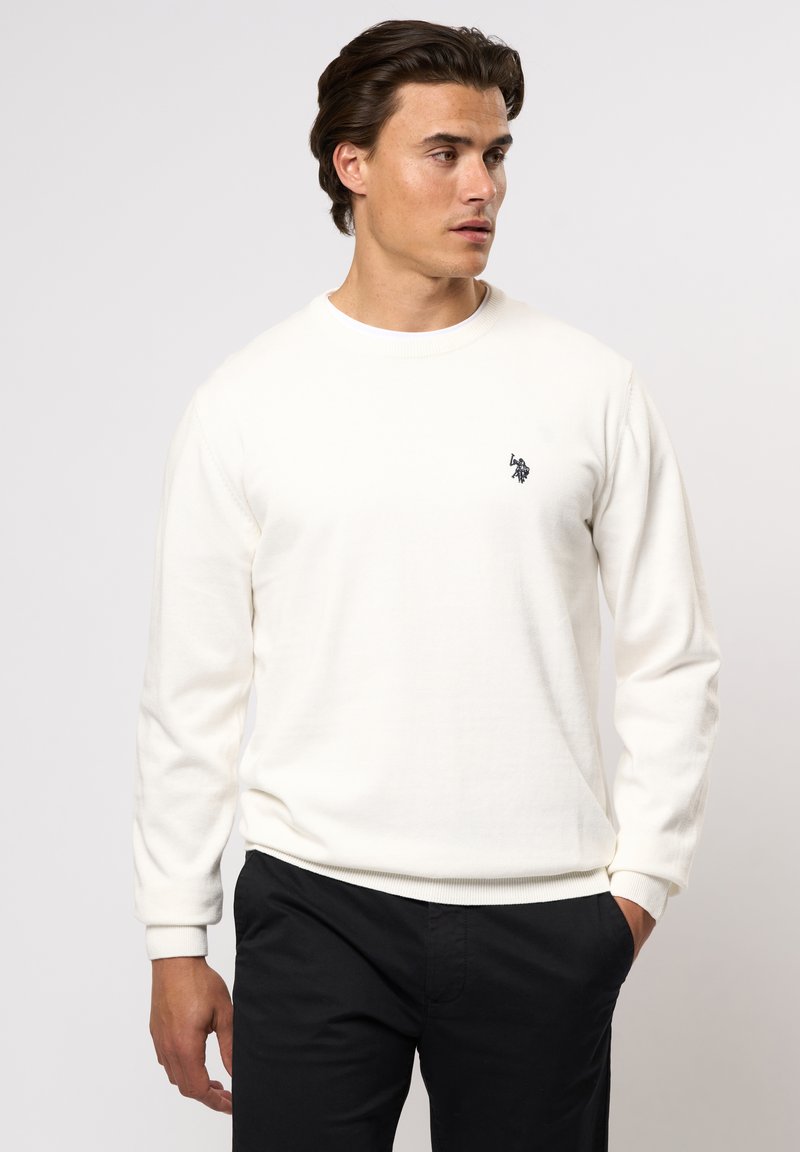 Sweatshirt blanc à manches longues en tissu doux, avec un col rond, des poignets et un ourlet côtelés, et un petit logo noir sur la poitrine gauche.