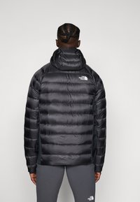 Schwarze gepolsterte Jacke mit Kapuze, ausgestattet mit horizontalen Steppmustern, dem The North Face-Logo im oberen Rückenbereich und einer glatten, glänzenden Oberfläche.