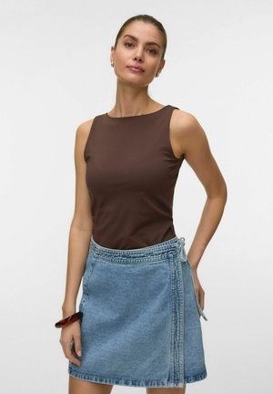 Young woman wearing a sleeveless brown top and light blue wrap denim skirt, standing against a plain white background.