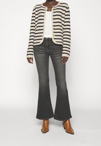 Raidallinen neulecardigan, jossa on kultaiset napit, yhdistettynä harmaisiin leveälahkeisiin farkkuihin ja ruskeisiin kärjiltään teräviin nilkkabootseihin. Valkoinen aluspaita näkyy päällä.