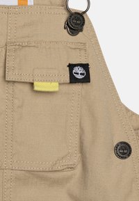 Salopette kaki fabriquée en tissu durable avec une poche avant, un onglet jaune et un patch logo de marque noir. Comprend des accents en métal.