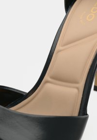 High-heeled black patent leather shoe with a beige cushioned insole featuring a smooth finish and elegant curve in the design.