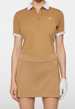 Frau trägt braunes kurzärmeliges Poloshirt mit weißem Kragen und passenden braunen Rock, hält weißen Golfhandschuh an der rechten Hand.