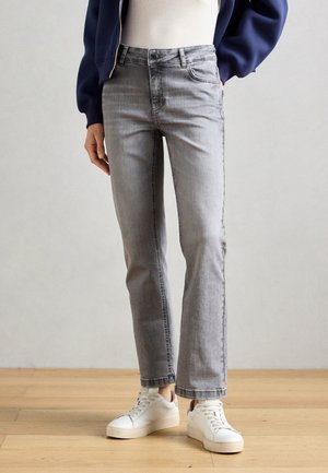 Person iført lysegrå jeans med lige ben, hvide sneakers, hvid top og navyblå jakke står på lyst trægulv.