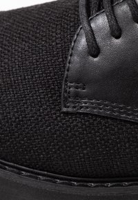 Chaussure noire avec une tige en tissu tissé et des accents en cuir lisse, dotée de lacets noirs et de coutures autour de la semelle en caoutchouc.