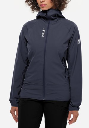 Frau trägt eine dunkelblaue Kapuzenjacke mit Reißverschluss und Millet-Logos auf Brust und Ärmel, kombiniert mit schwarzen Hosen, steht vor weißem Hintergrund.