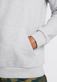 La mano derecha descansando dentro del bolsillo delantero de una sudadera gris claro, combinada con pantalones de camuflaje verde.
