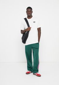 Hvid kortærmet t-shirt med sort logo, grøn sweatpants med pink striber, og røde sko. Sort skuldertaske hængt over kroppen.