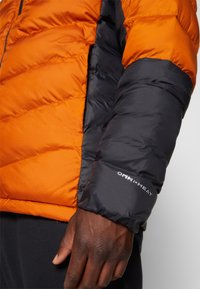 Detailní záběr na ruku osoby v oranžové a černé péřové bundě, na které se nachází logo "OMNI-HEAT" na rukávu.