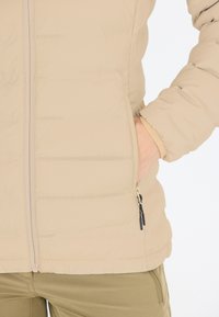 Beige puffjacka med dragkedja fram, lätt material, horisontell sömnad och sidofickor med svarta dragkedjehandtag.