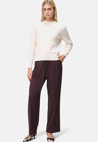 Cremefarbener, gestrickter Pullover mit durchbrochenem Muster und gewelltem Ausschnitt, kombiniert mit dunkelbraunen, weit geschnittenen Hosen und gemusterten Schuhen.