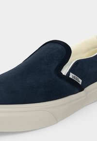 Μπλε σκούρα Vans slip-on παπούτσια με λευκή λαστιχένια σόλα και μικρή ετικέτα Vans στο πλάι, εμφανιζόμενα σε απλό ανοιχτό φόντο.
