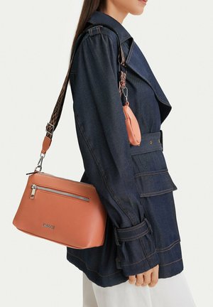 Žena v tmavě modré džínové bundě nesoucí středně hnědou koženou kabelku přes rameno s předním zipem a vzorovaným nastavitelným popruhem.